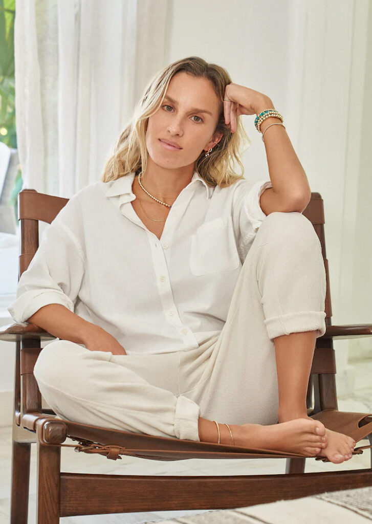 Alexa Leigh sits barefoot on a wooden chair, dressed in a relaxed white outfit and adorned with her signature jewelry